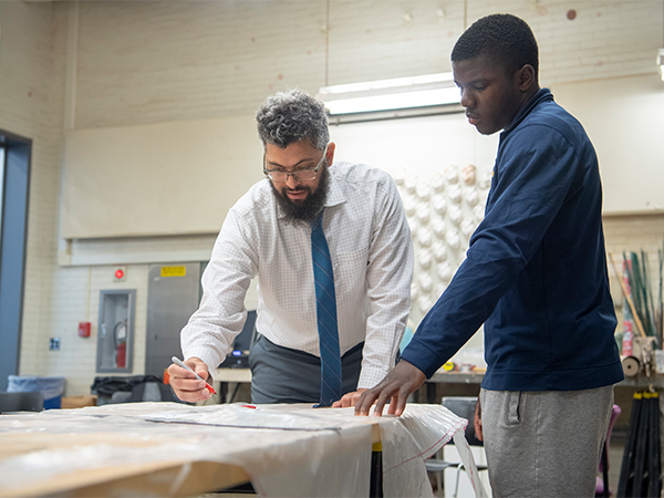 A male instructor works on a piece of art in a studio as a student looks on.